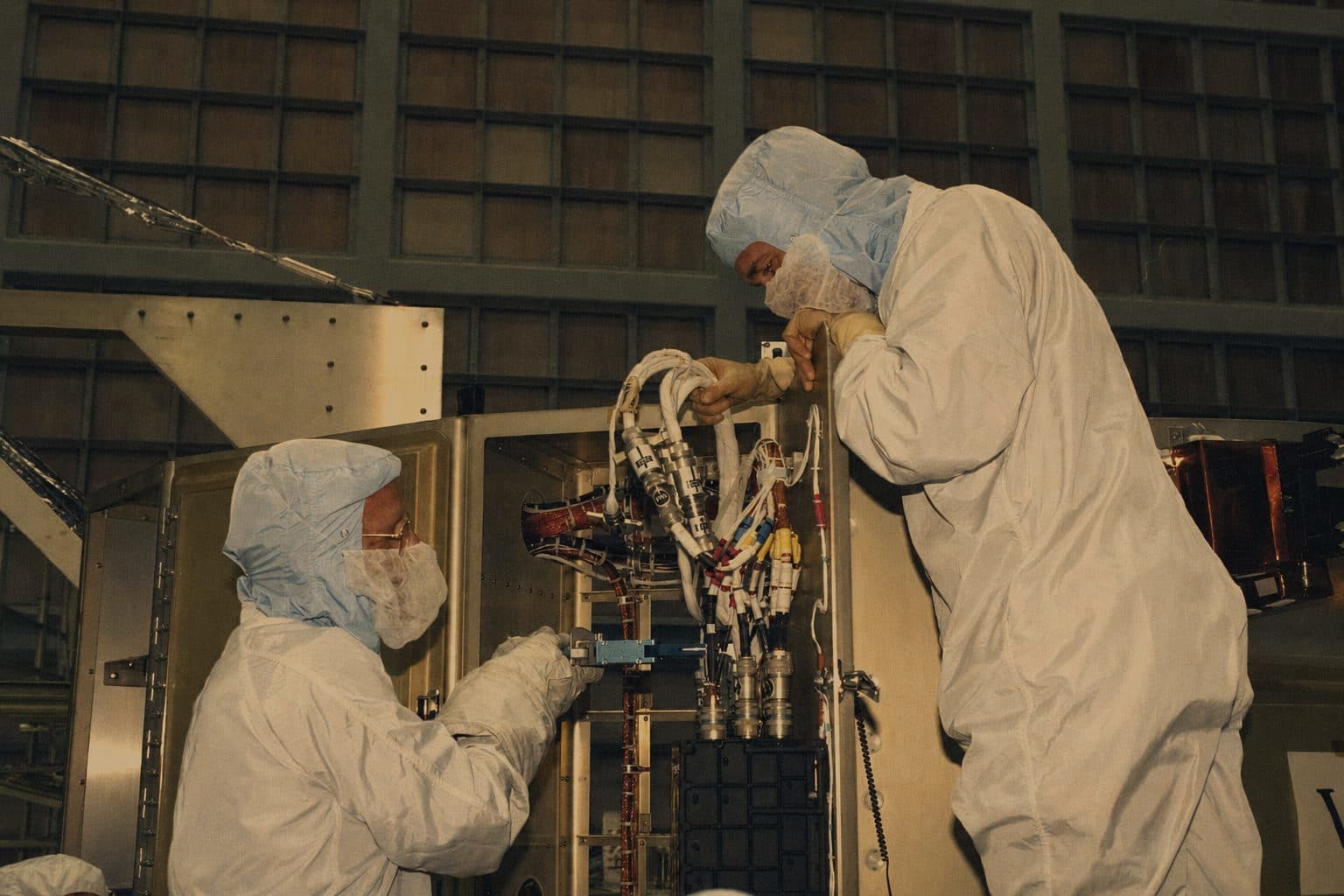 Two engineers in cleanroom suits working on flight hardware inside an assembly bay, one steadying a fitting while the other reaches into the structure.