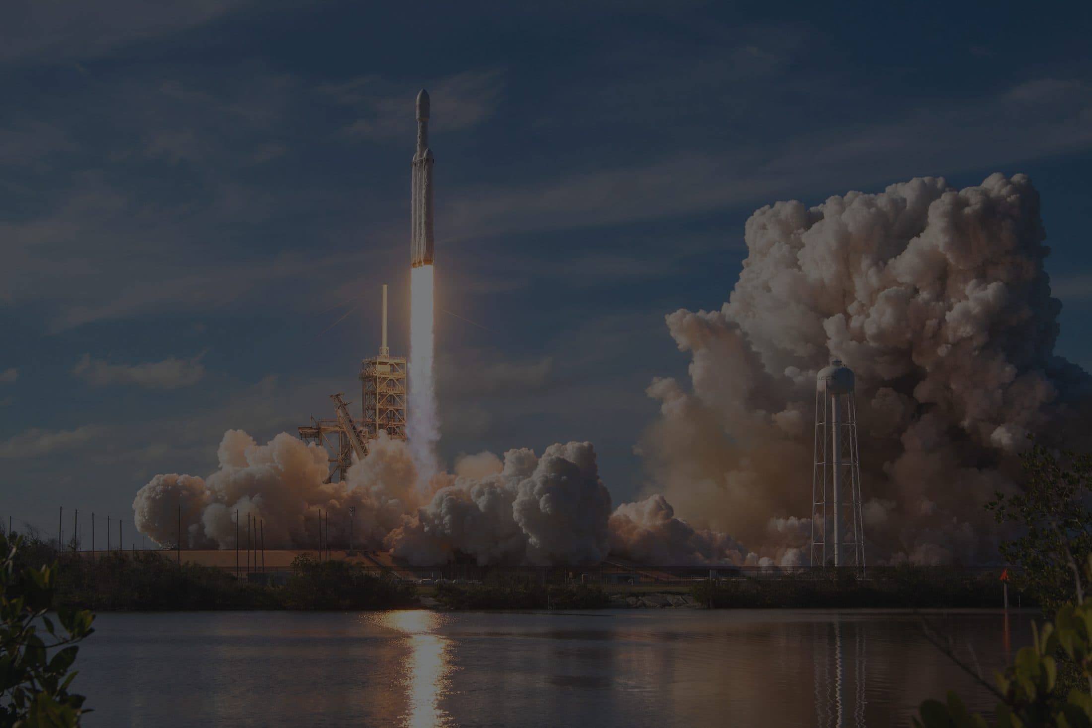 A Falcon Heavy lifting off its pad, the vehicle climbing on a single bright column of flame with the exhaust cloud rolling out across the ground and reflecting in the water in front of it.