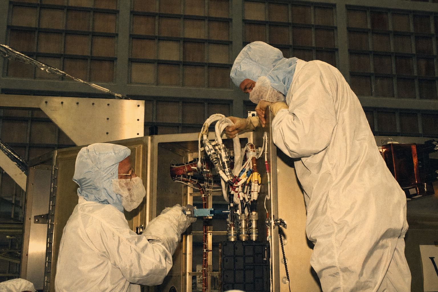 Two technicians in cleanroom suits connecting a wiring harness inside a spacecraft structure.