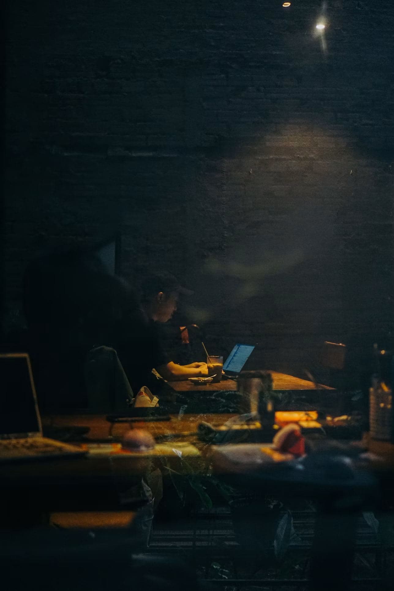 An engineer working alone at a laptop late at night, seen through a window.