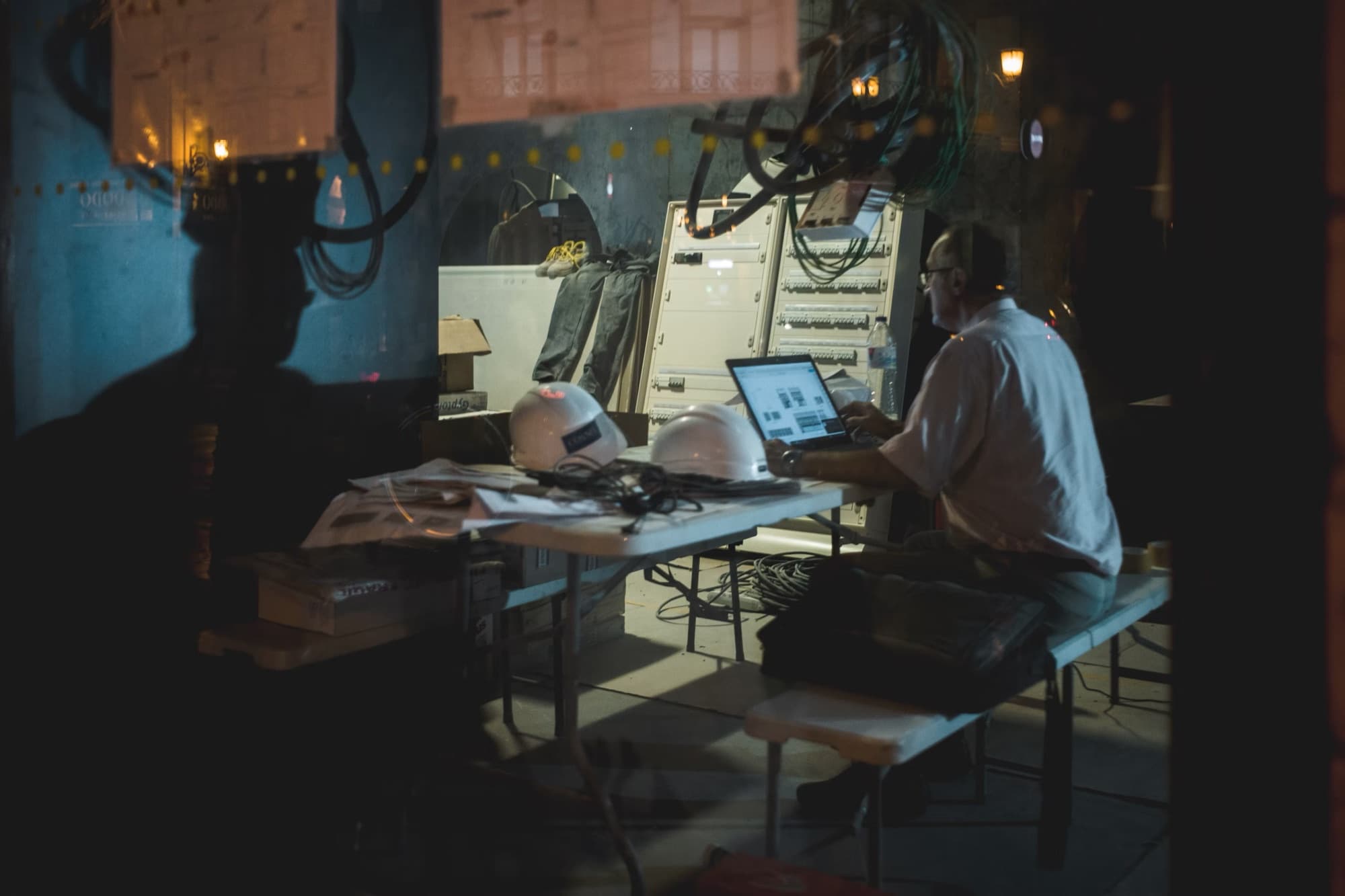 An engineer working late over a laptop on site, hard hats and an open control cabinet beside them.