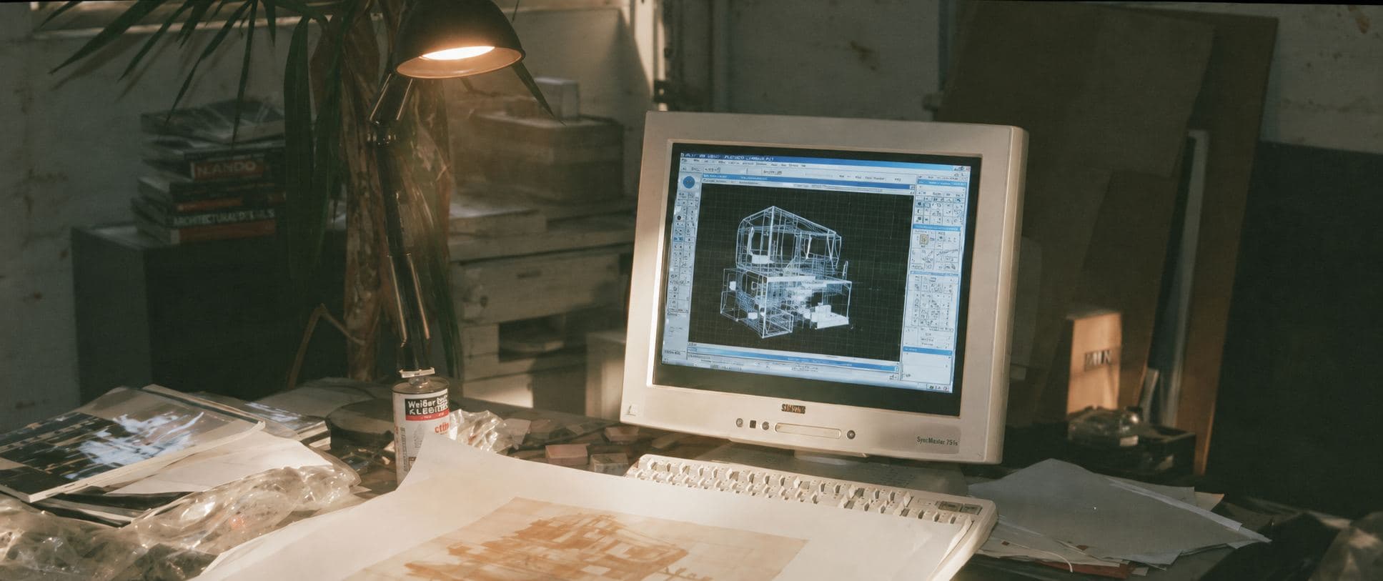 A CRT monitor on a cluttered studio desk at golden hour, running early CAD software with a wireframe model on screen, a drawing lying in front of it.