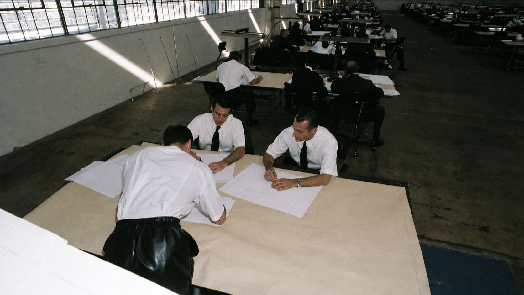 A vast mid-century drafting office: rows of drafting tables under factory windows, men in white shirts and ties leaning over drawings.
