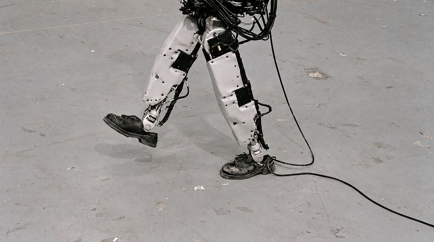 The legs of a humanoid robot mid-stride across a concrete floor, wearing scuffed leather work boots, trailing a cable.