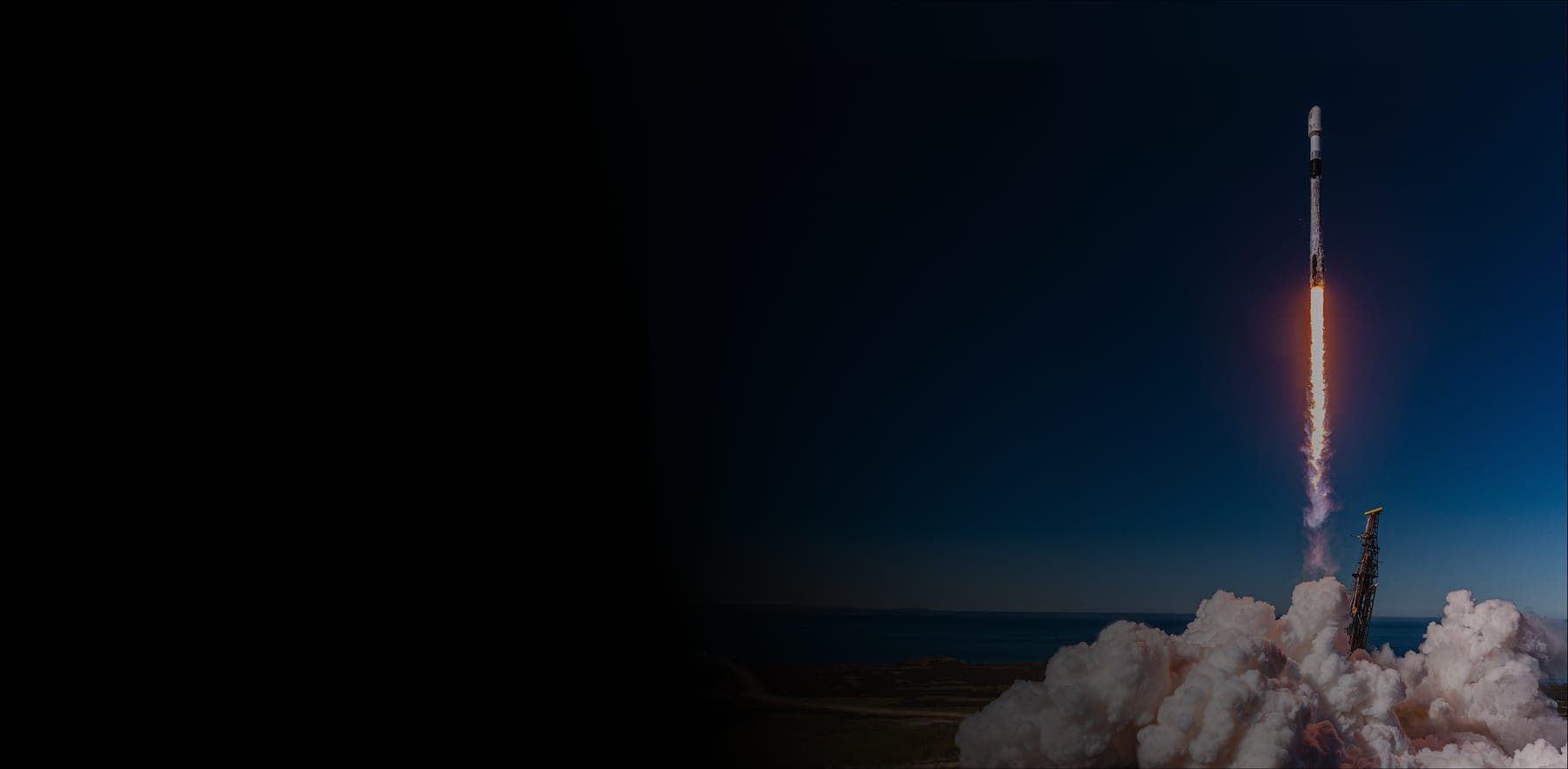 A Falcon 9 rocket climbing away from its pad at dusk, exhaust lit against a deep blue sky over the ocean.
