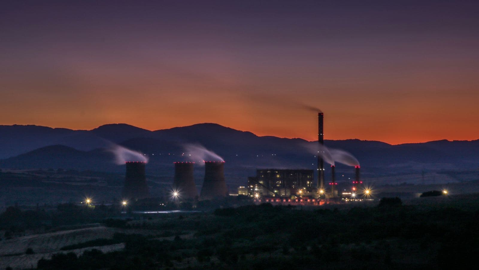 A power station at dusk, its cooling towers venting steam against a ridge line with the last of the sunset behind it.