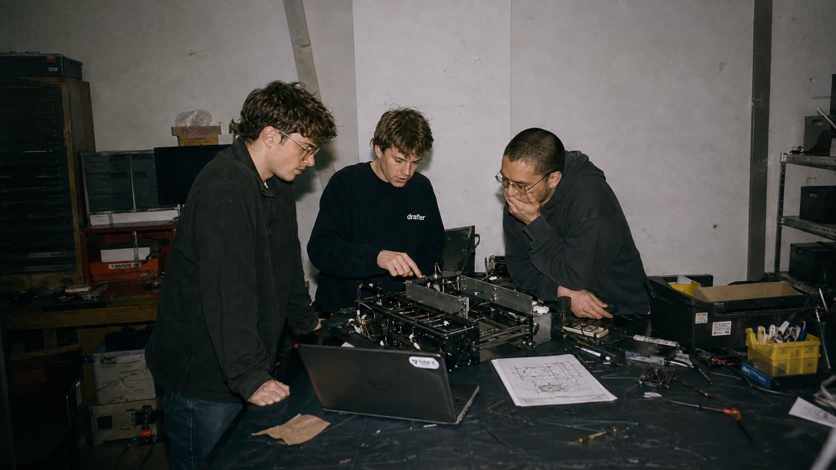 Three Drafter engineers leaning over a machined fixture on a workshop bench, a laptop and a printed drawing beside it, one of them wearing the Drafter crewneck.