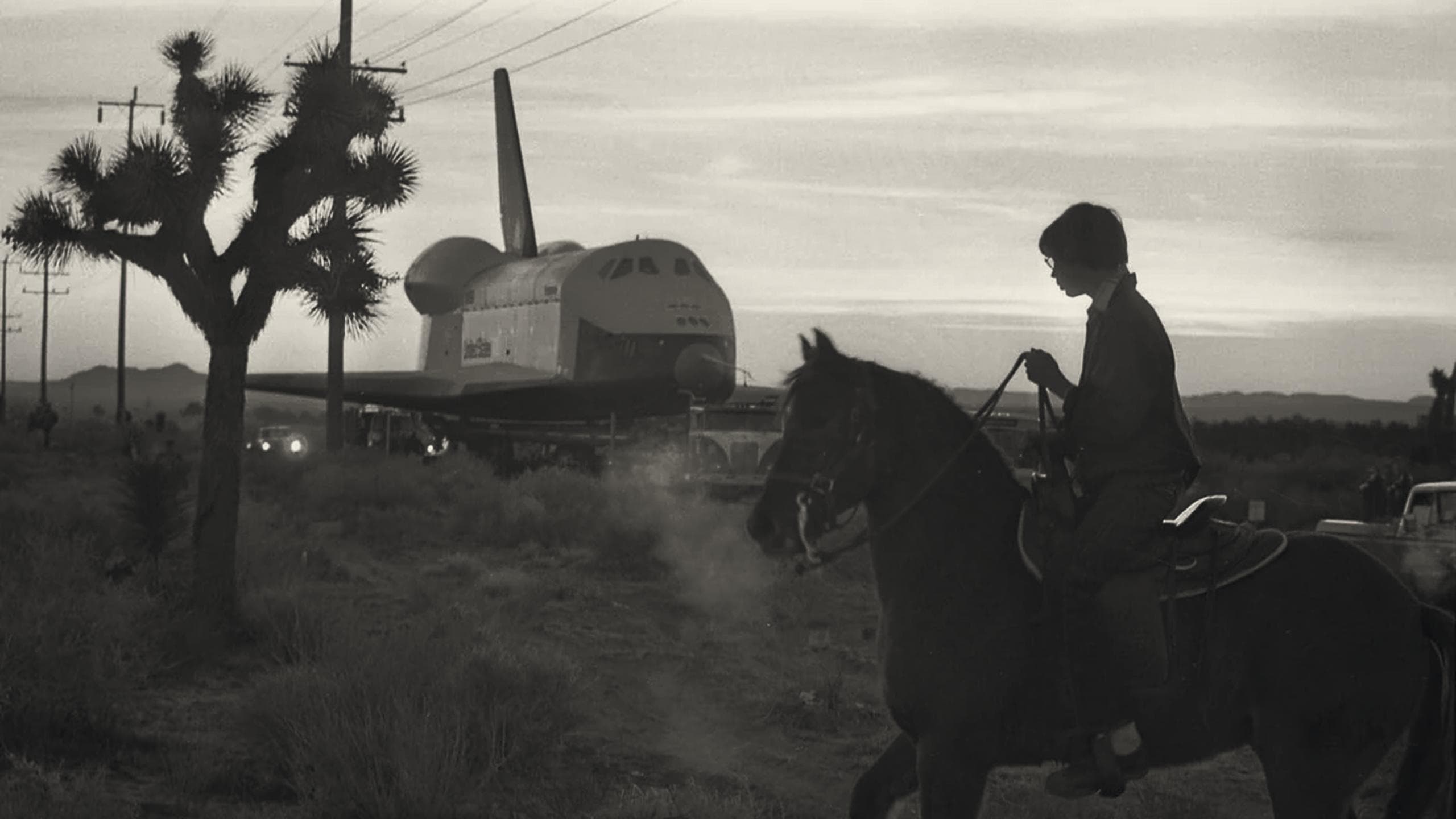 A rider on horseback at dusk, watching the Space Shuttle Enterprise being towed down a desert road past Joshua trees.